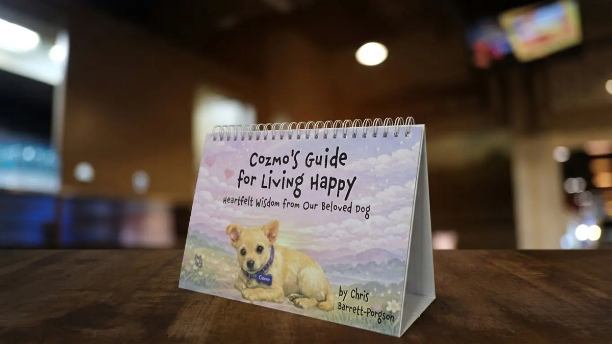 A standing desk calendar display of Cozmo's Guide for Living Happy: Heartfelt Wisdom from Our Beloved Dog by Chris Barrett-Forgach, featuring an illustration of Cozmo on the cover, set on a wooden table